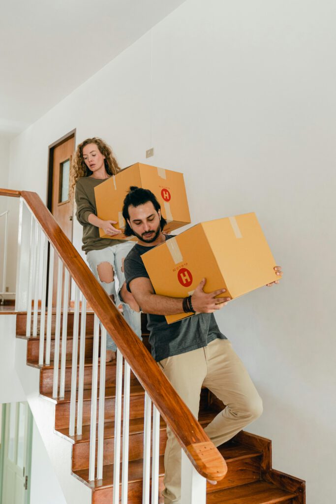 A young couple carrying boxes down stairs in a modern home, indicating a move or relocation.